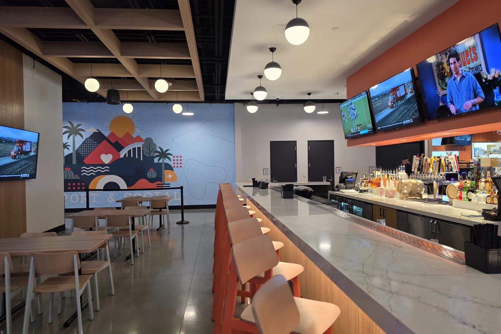 The bar at Henderson Sport & Social, with a long white counter, orange-backed stools, sports on a row of TVs above, and a mountain mural on the far wall of the dining area