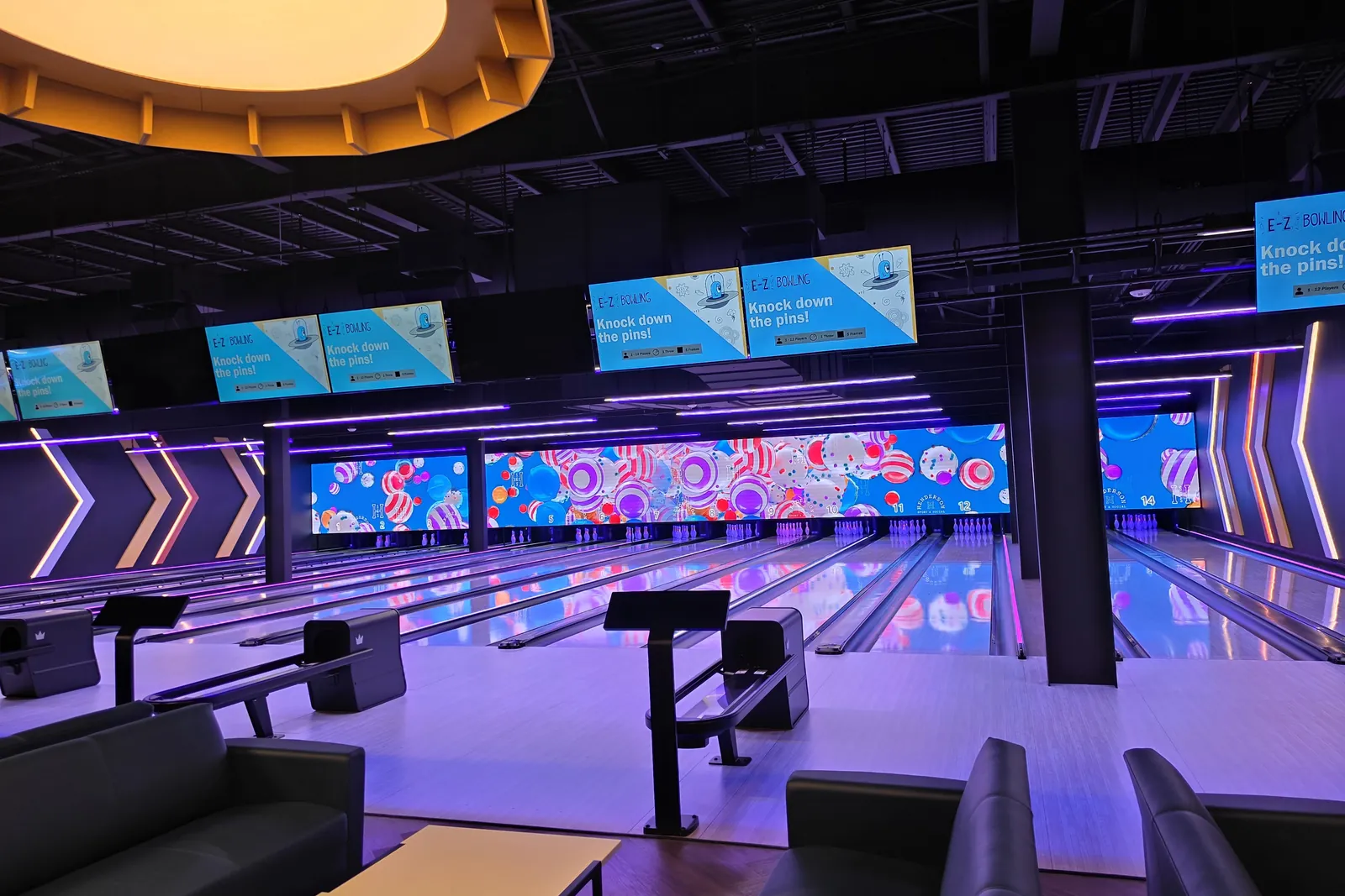 Main bowling lanes at Henderson Sport & Social under purple lighting, with colorful LED screens above the pins, overhead score monitors and leather lounge seating in the foreground
