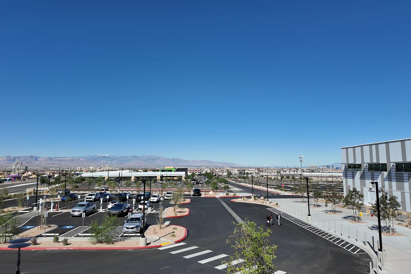 Elevated view from the Henderson Sport & Social balcony over its parking lot and the gray-striped side of the building, with West Henderson development and mountains under a clear blue sky