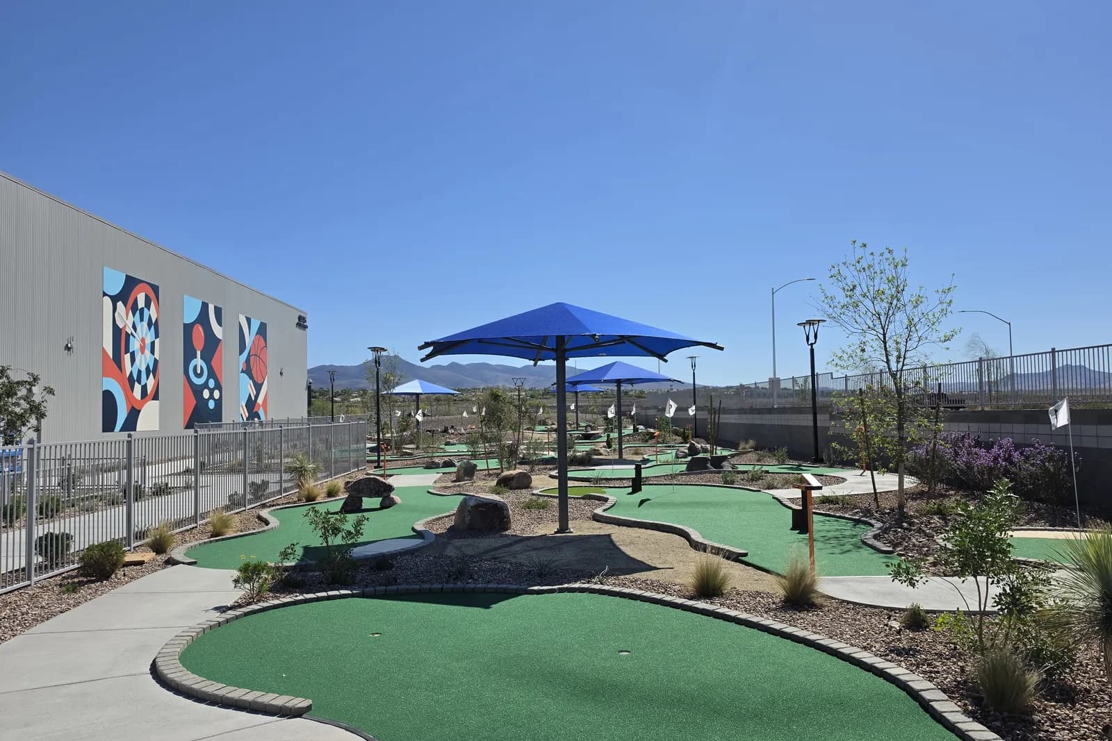 Outdoor 18-hole mini golf course beside the Henderson Sport & Social building, with green turf, blue shade umbrellas, desert landscaping and a colorful mural on the wall