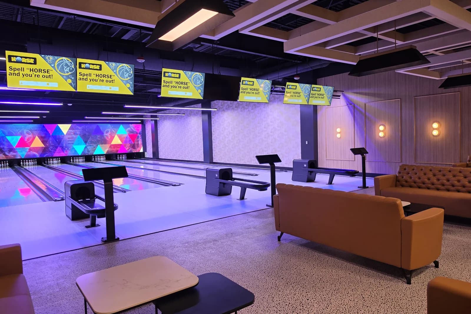 The VIP bowling room at Henderson Sport & Social, with tan upholstered sofas and small tables facing a row of lanes under a geometric LED wall