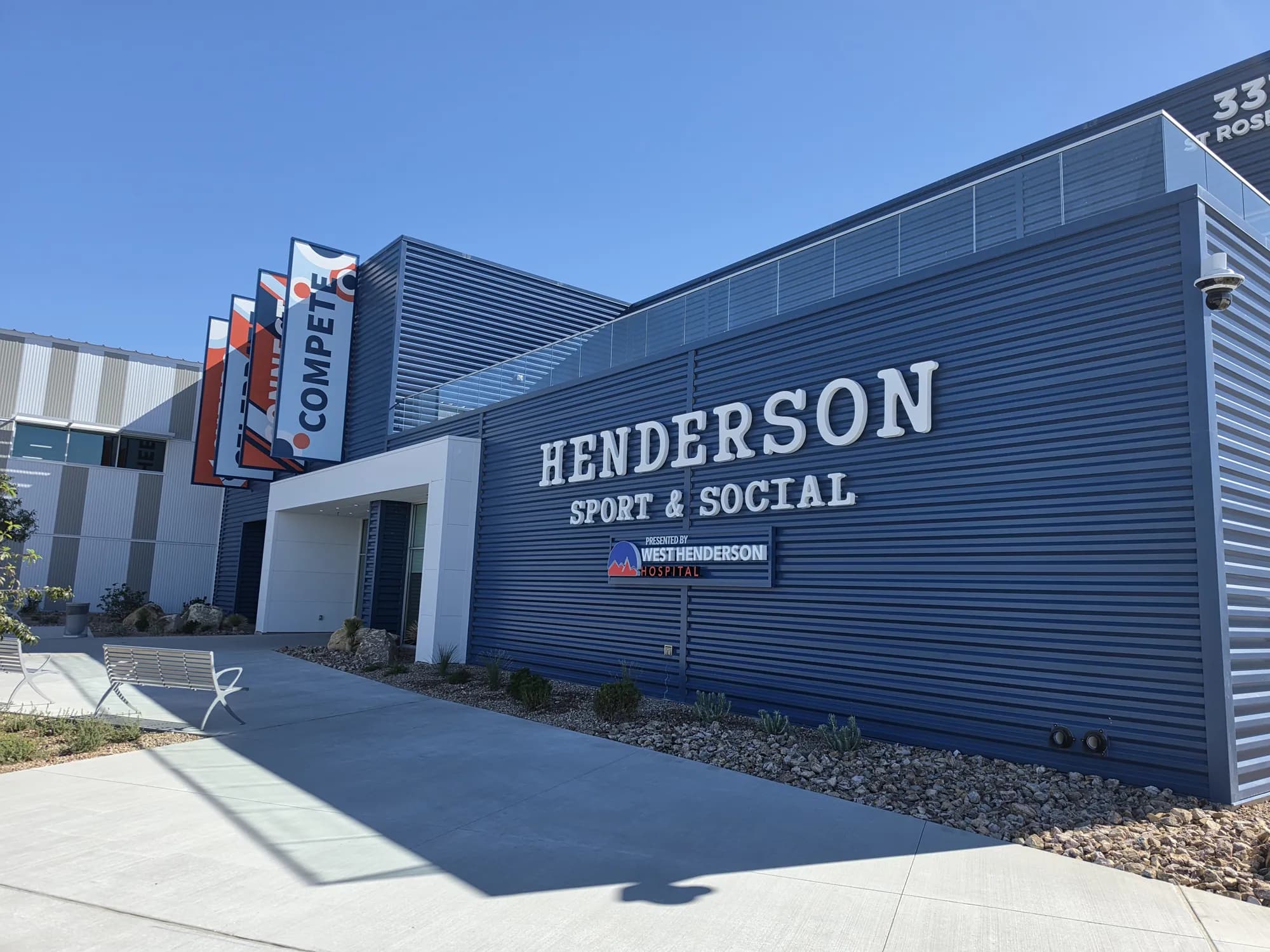 The navy-blue entrance wing of Henderson Sport & Social with large HENDERSON SPORT & SOCIAL lettering, the 3375 St. Rose Pkwy address and a COMPETE banner, under a clear blue sky in West Henderson