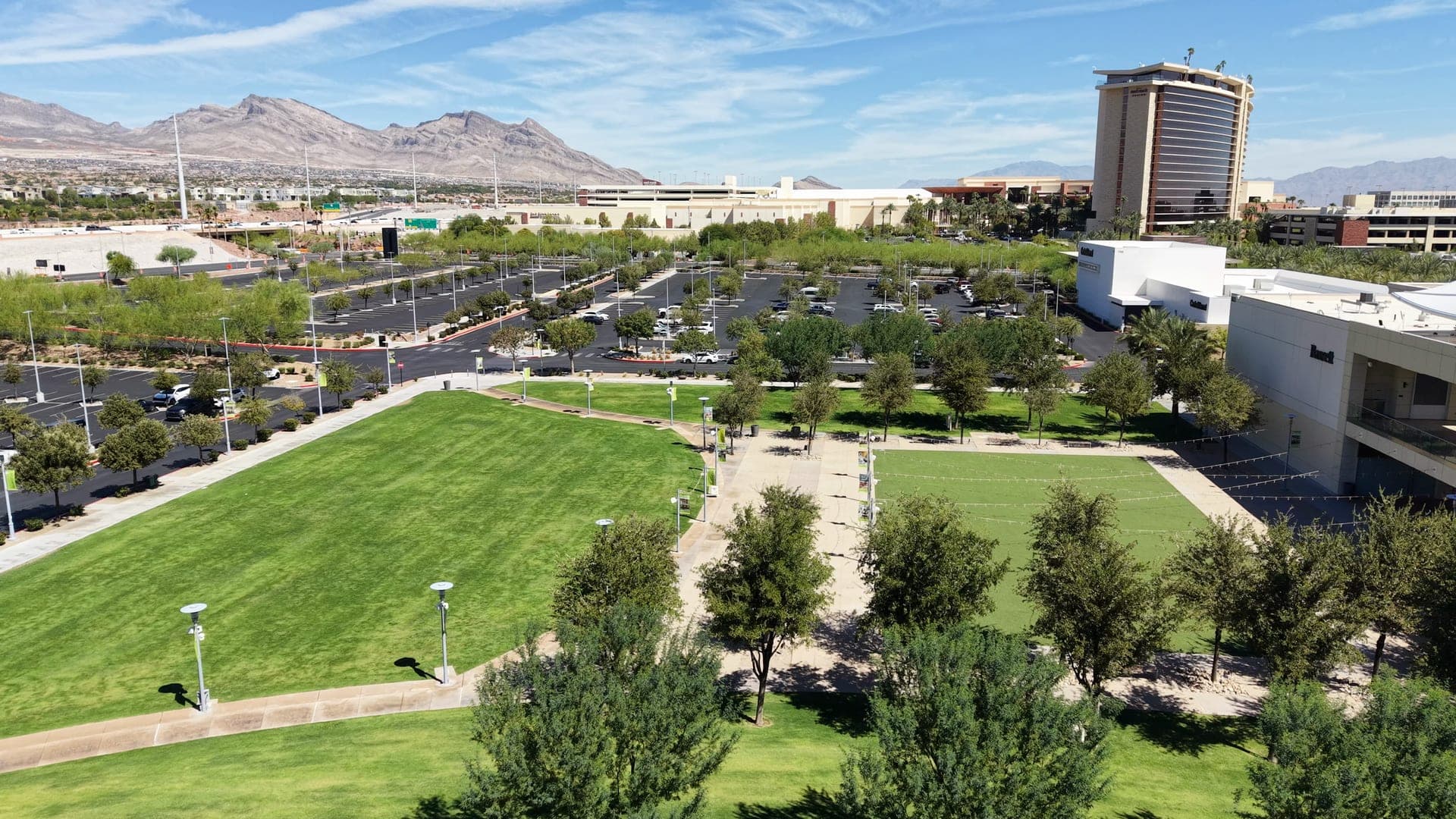 Aerial view of Downtown Summerlin's green lawns and tree-lined walkways beside retail buildings, with a casino hotel tower, parking areas and desert mountains behind.