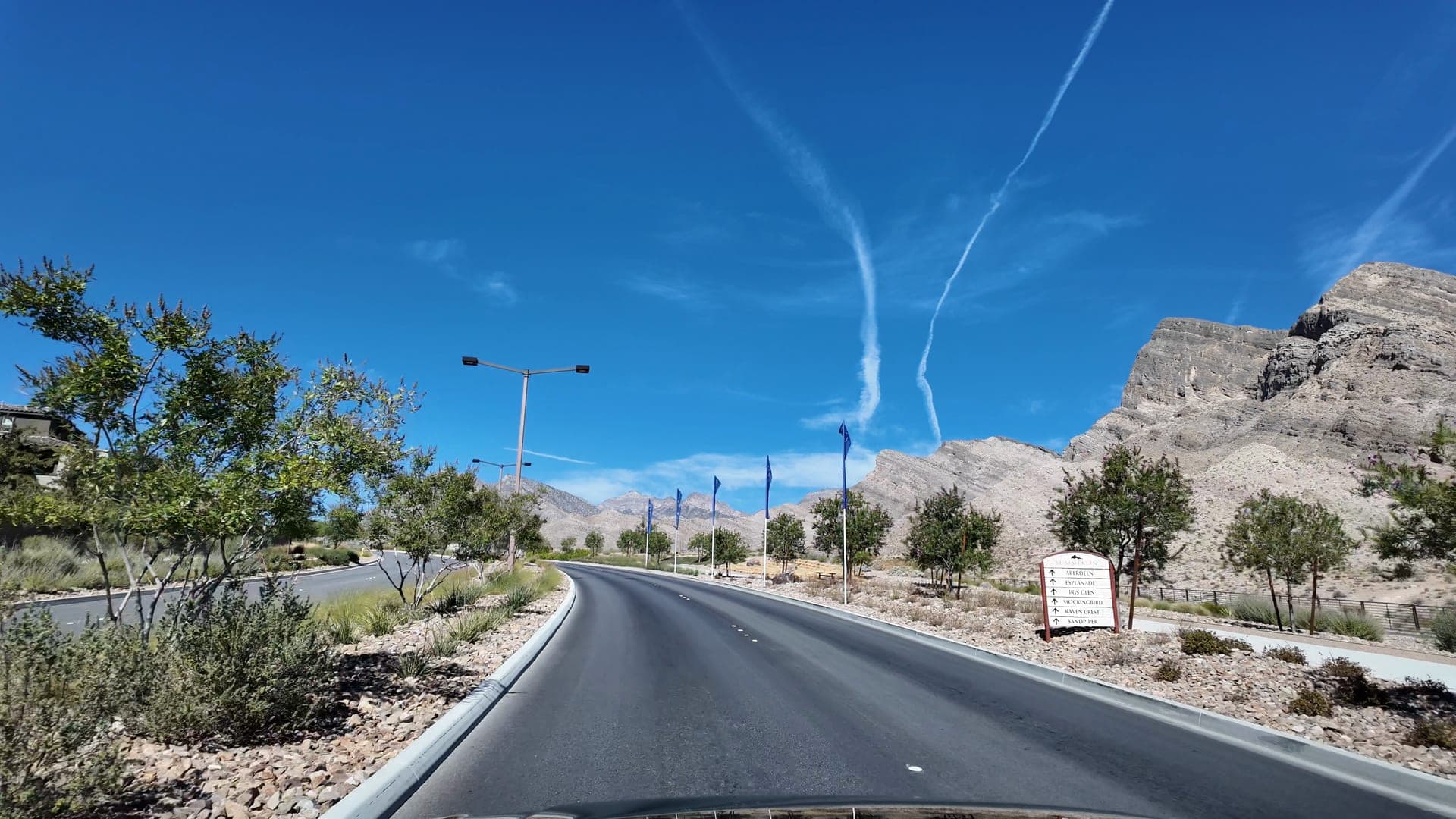 Curving landscaped road in Summerlin with blue banner flags and a neighborhood directional sign, running toward tall sandstone cliffs.