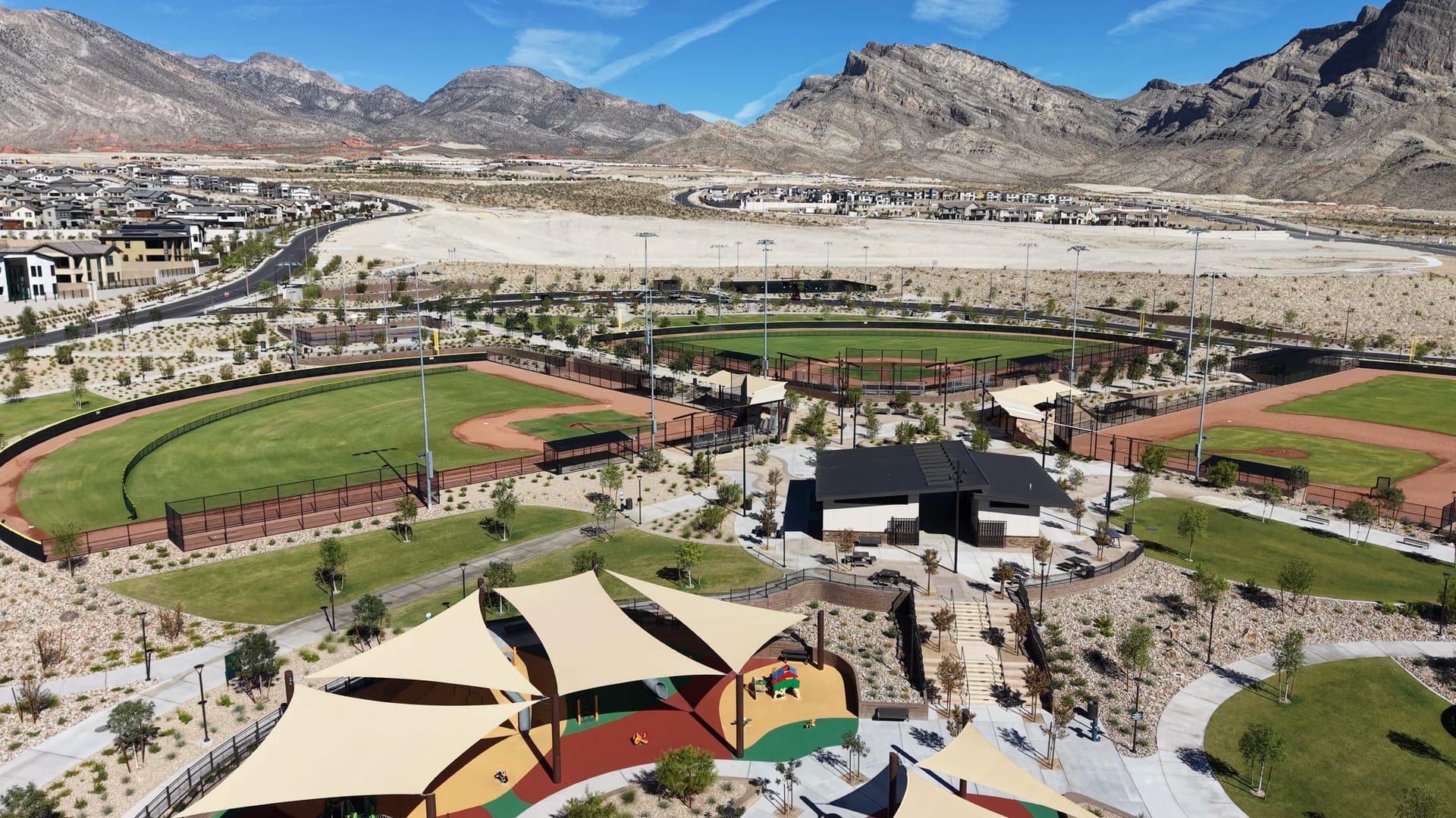 Aerial view of Grand Park in Summerlin West: a shade-sail playground and lit ballfields in the foreground, with new home neighborhoods, graded land and rugged mountains behind.