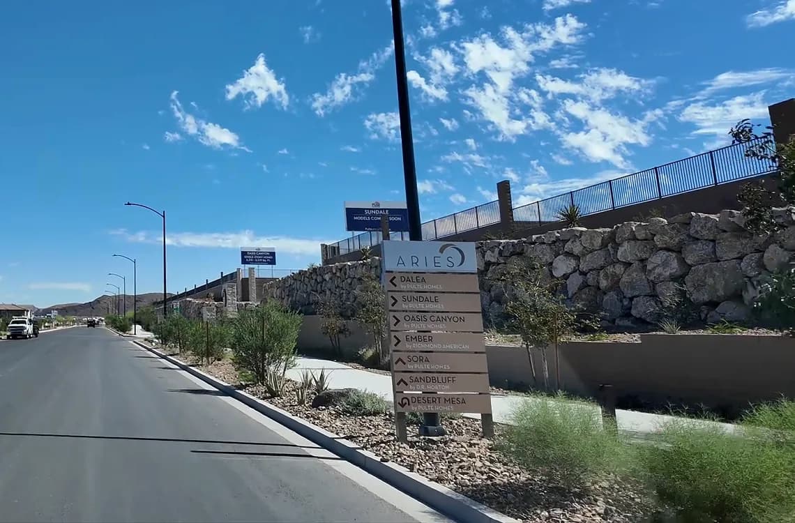 Aries wayfinding sign beside a community road listing Dalea, Sundale, Oasis Canyon, Sora and Desert Mesa by Pulte Homes, Ember by Richmond American and Sandbluff by D.R. Horton, with a Sundale models coming soon billboard behind it.