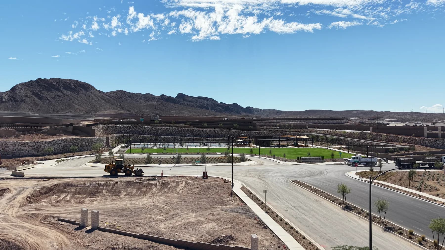 Aerial view of Greenbow Park at Aries, with green turf, shade structures and terraced rock retaining walls, and a loader, trucks and graded dirt in the foreground.