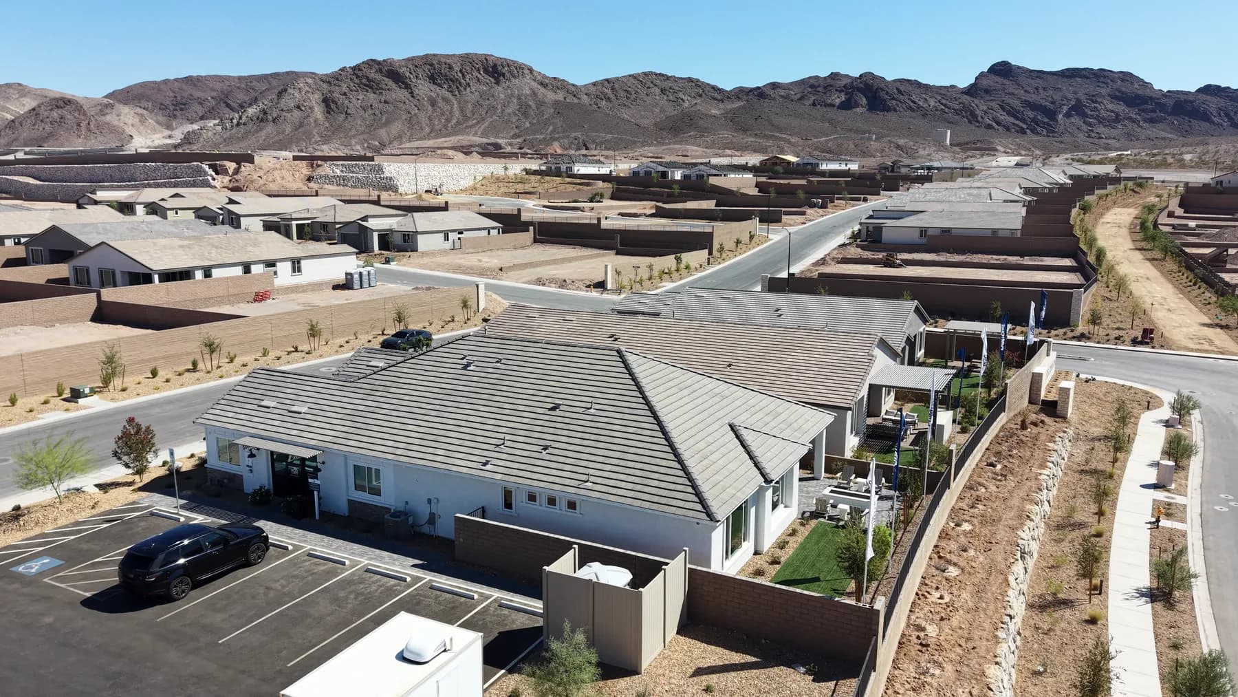 Aerial view over a single-story model home and its parking lot in Pulte's Oasis Canyon at Aries, with finished homes, walled empty lots, new streets and desert mountains behind.
