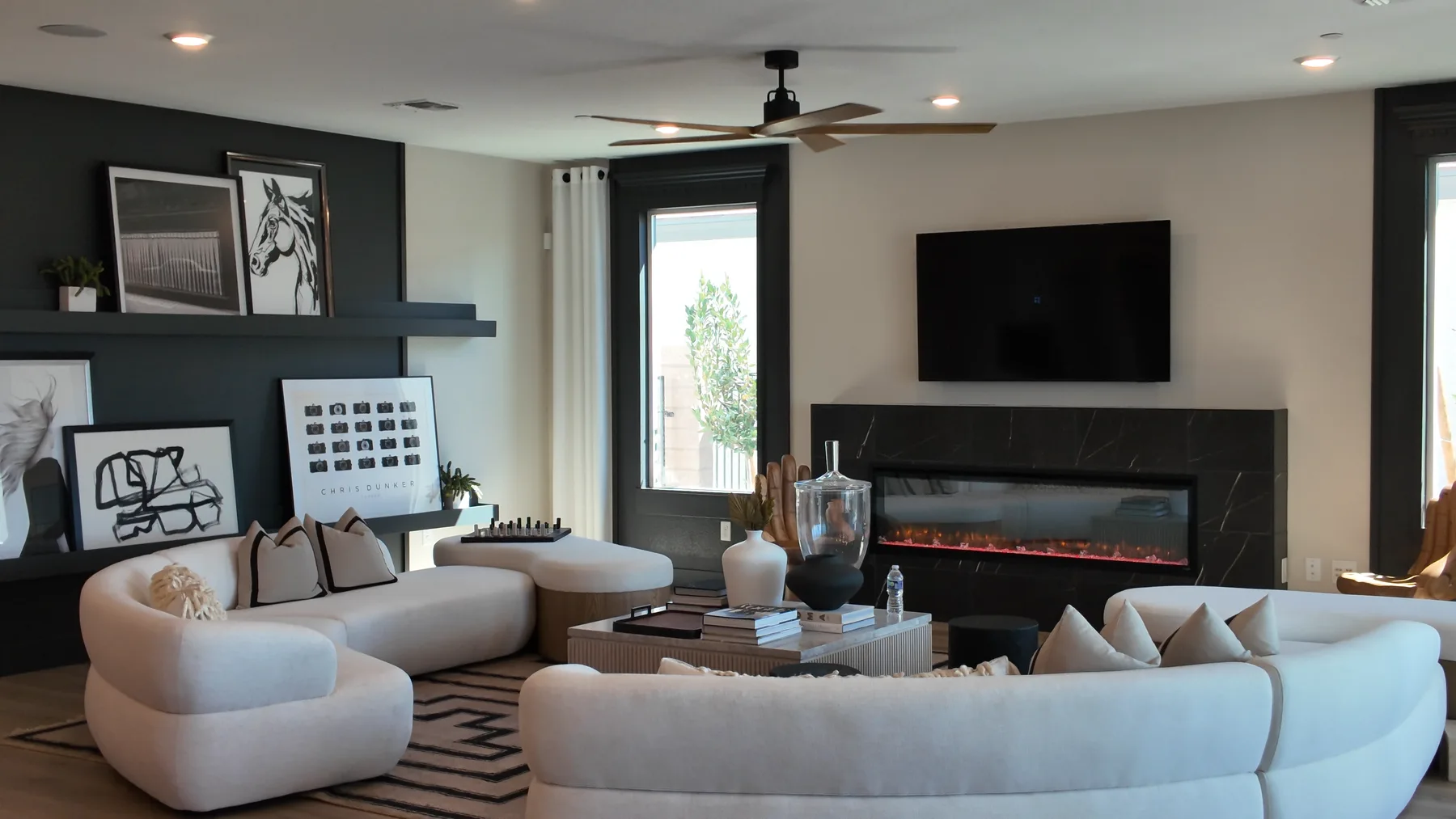 Living room of Pulte's Parklane model at Oasis Canyon in Aries, with curved white sofas, a black stone fireplace wall with a linear electric fireplace, a wall-mounted TV and framed black-and-white art.