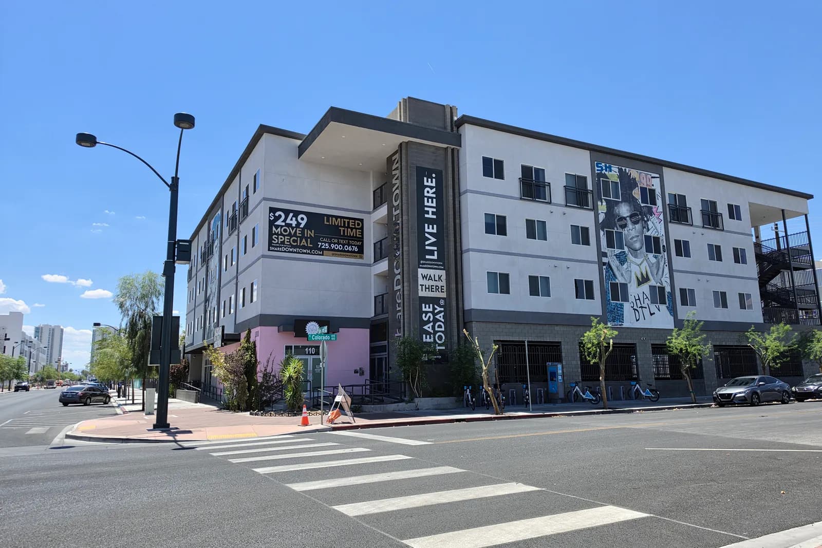 A newly built four-story apartment building on the edge of the Las Vegas Arts District with a large street-art mural, bike-share docks out front, and signage reading Live Here, Walk There.