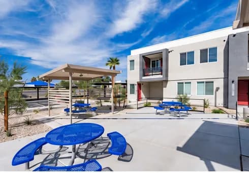 A courtyard at Gholson Landing with blue picnic tables under a shade structure, next to one of the apartment buildings