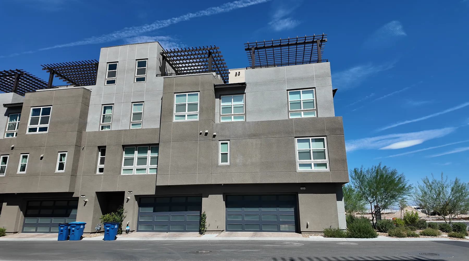 Street-level view of newly built three-story townhomes in Inspirada, Henderson, with flat-roofed modern massing, rooftop pergolas, ground-floor garages and young desert landscaping along the curb.