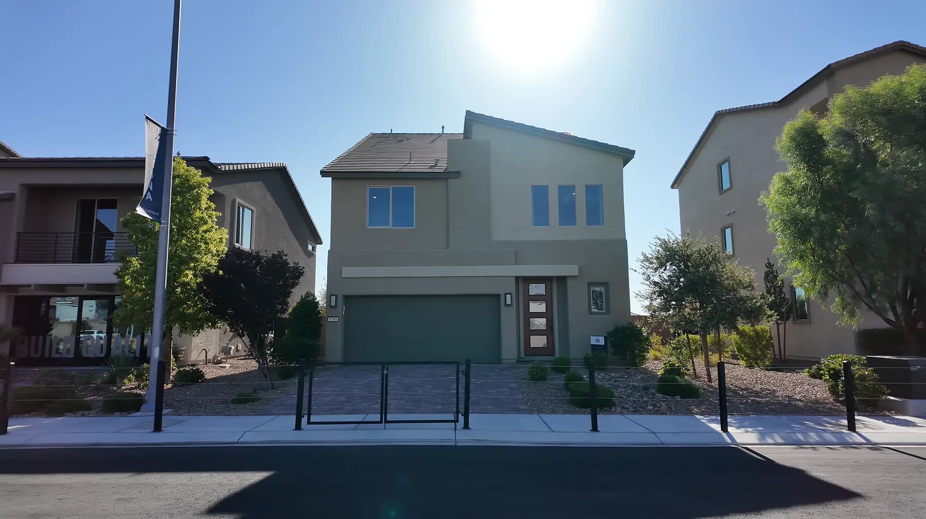 A two-story new-construction model home in southwest Las Vegas with a builder flag on a pole out front, a low metal rail along the sidewalk, young shrubs in fresh rock landscaping, and neighboring new homes on either side.