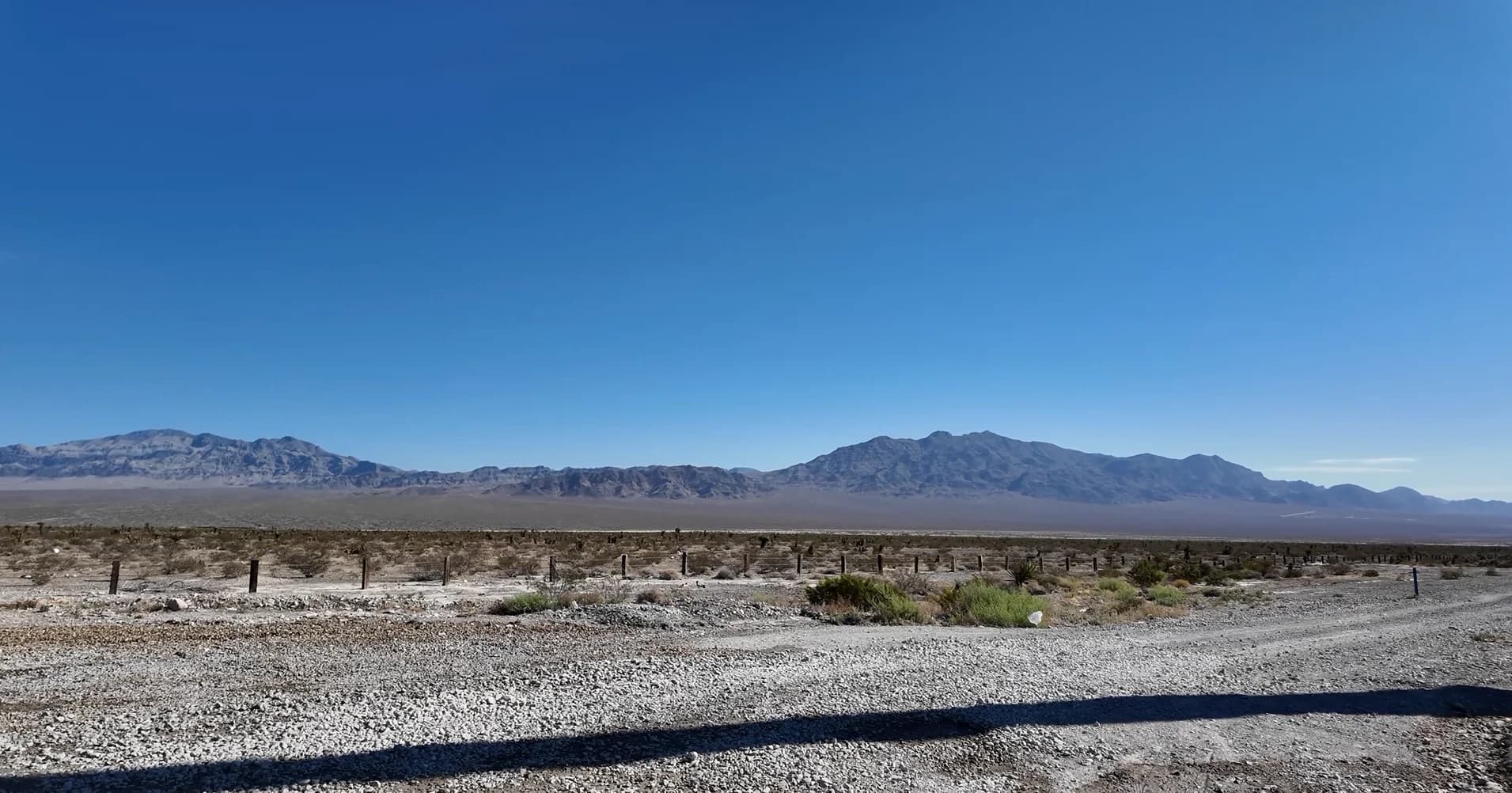 Ground-level view of the vacant Monument Hills desert site, looking toward the Spring Mountains, with a fence line and gravel road in the foreground