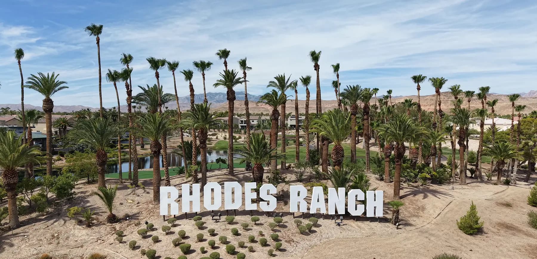 Aerial drone view of the Rhodes Ranch entry sign in southwest Las Vegas, backed by tall mature palms, a green golf corridor with water features, and homes among grown-in landscaping with mountains on the horizon.