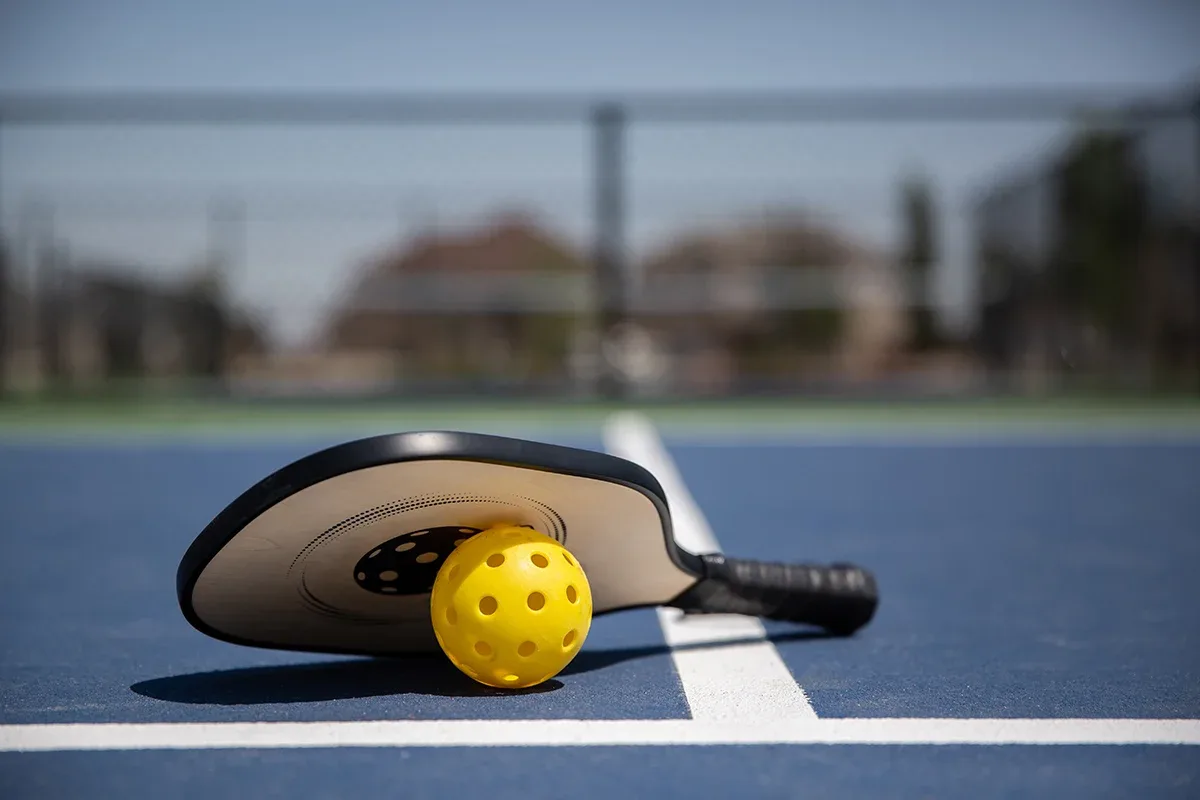 A pickleball paddle and ball resting on a court, with houses visible in the background