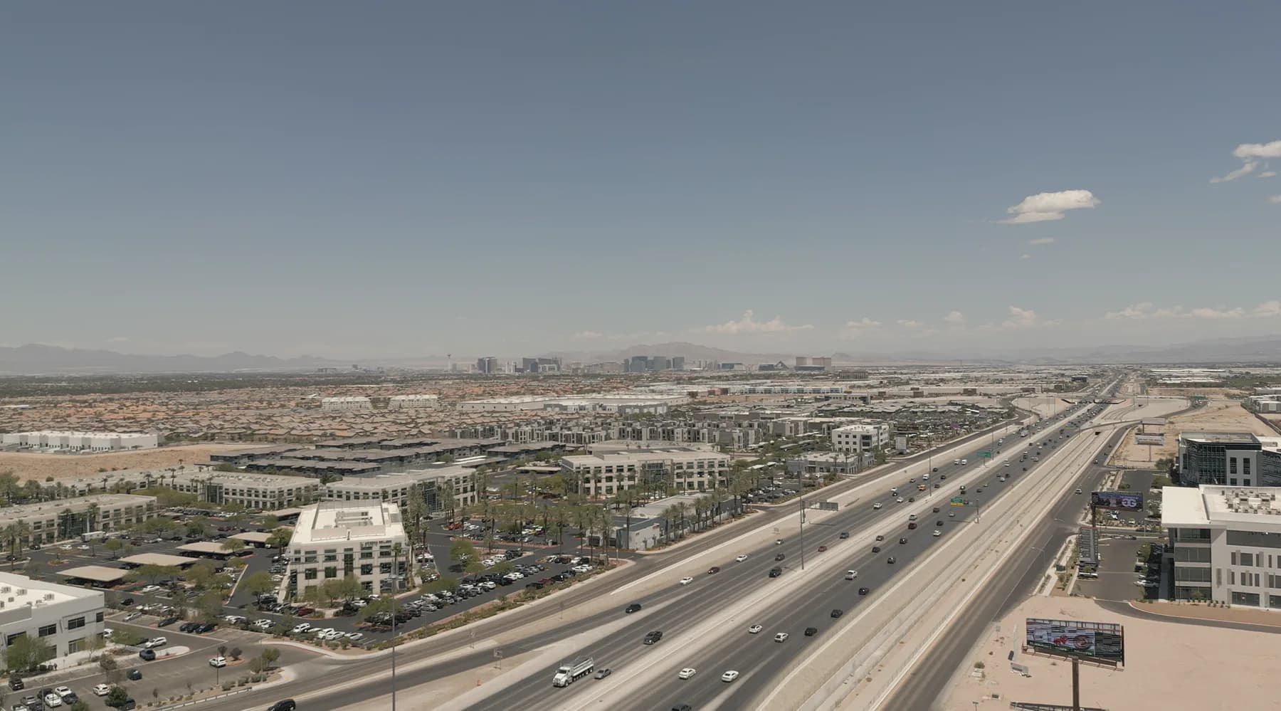 Aerial view along the 215 beltway through southwest Las Vegas, offices and rooftops on both sides of the freeway and the Strip skyline on the horizon.