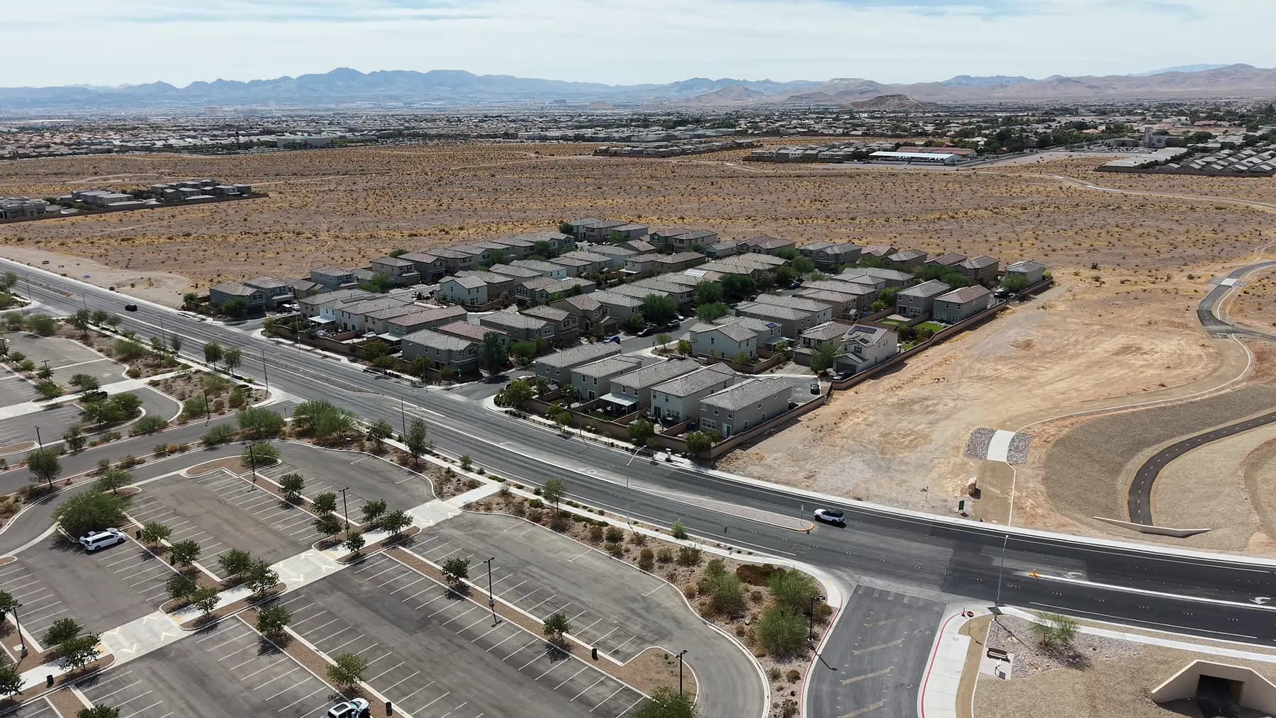 Aerial view in southwest Las Vegas of a finished block of homes ending abruptly at open, undeveloped desert, with a newly built road and an empty parking lot alongside.