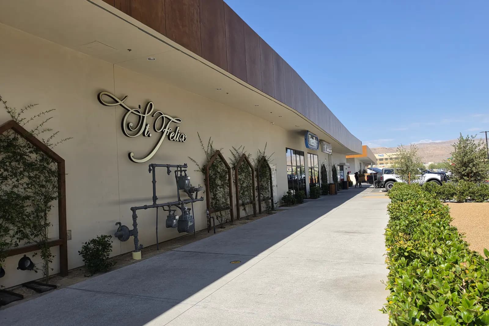 The sidewalk along a row of storefronts at The Bend in southwest Las Vegas, with St. Felix and Freed's signage, planted trellises against a stucco wall, trimmed hedges, parked trucks, and desert hills on the horizon.