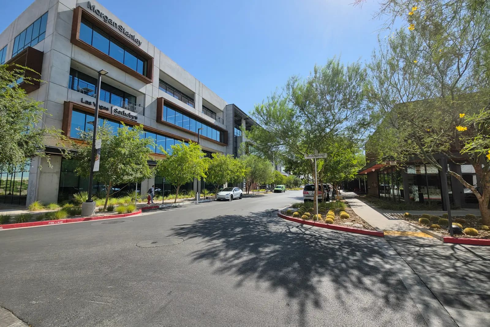 A mid-morning street at the UnCommons campus in southwest Las Vegas, with a four-story office building signed for Morgan Stanley and Las Vegas Sotheby's International Realty, parked cars, and young trees casting shadows across the asphalt.
