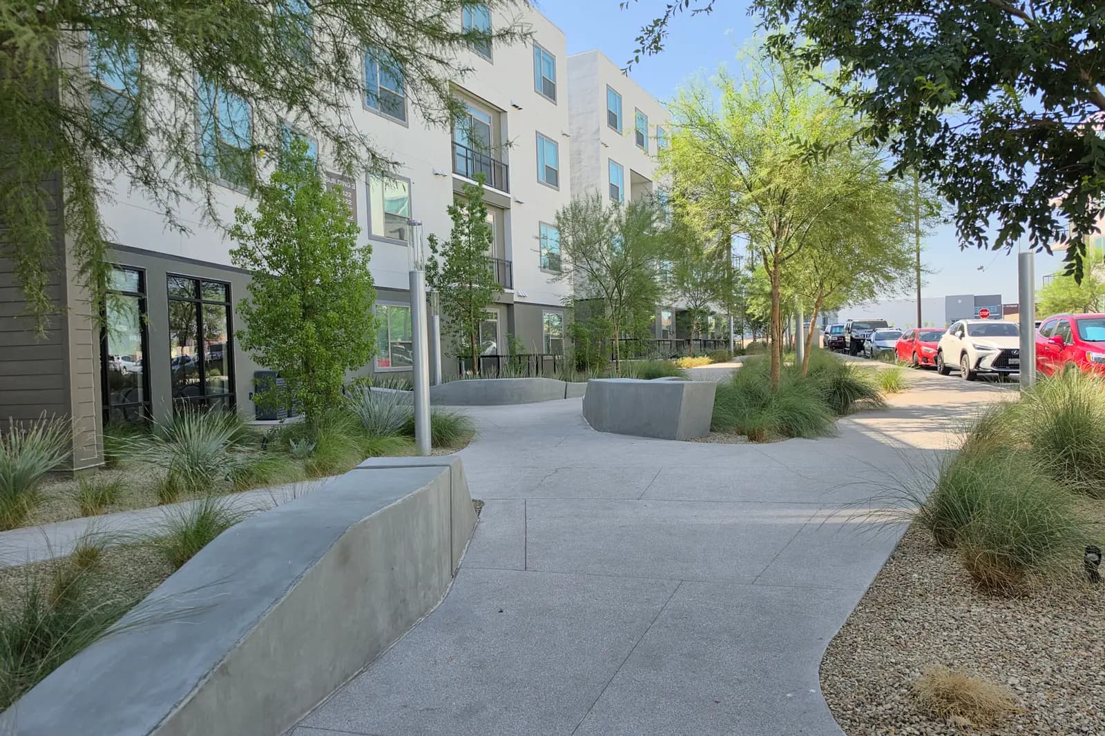 A shaded concrete walkway between apartment buildings at UnCommons in southwest Las Vegas, with low concrete seating walls, desert grasses, young trees, and parked cars visible at the far end.