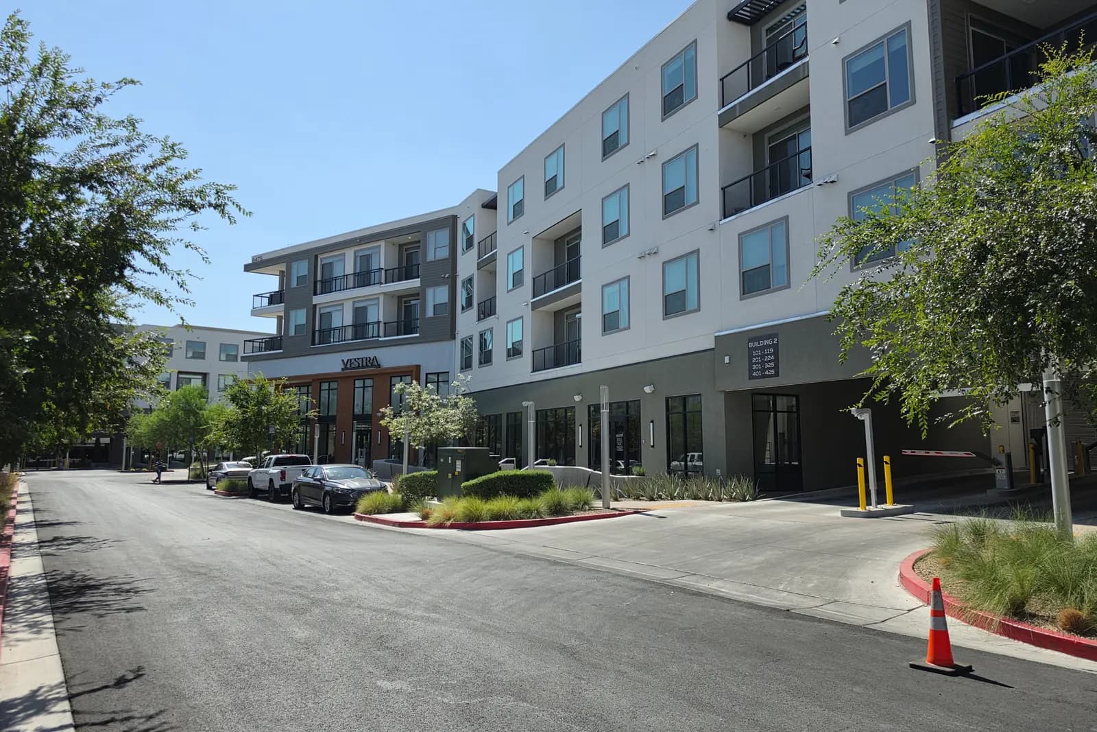 The four-story Vestra apartment building at UnCommons in southwest Las Vegas, with balconies, ground-floor retail bays, a leasing entrance, and cars parked along the street on a clear July morning.