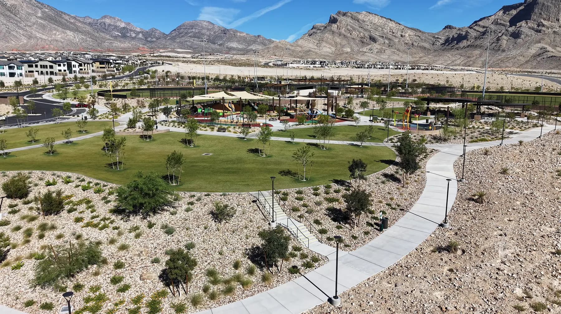 Aerial view of Grand Park in Summerlin West, a green lawn and shaded playground with walking paths, new rooftops on the left and the mountains behind.