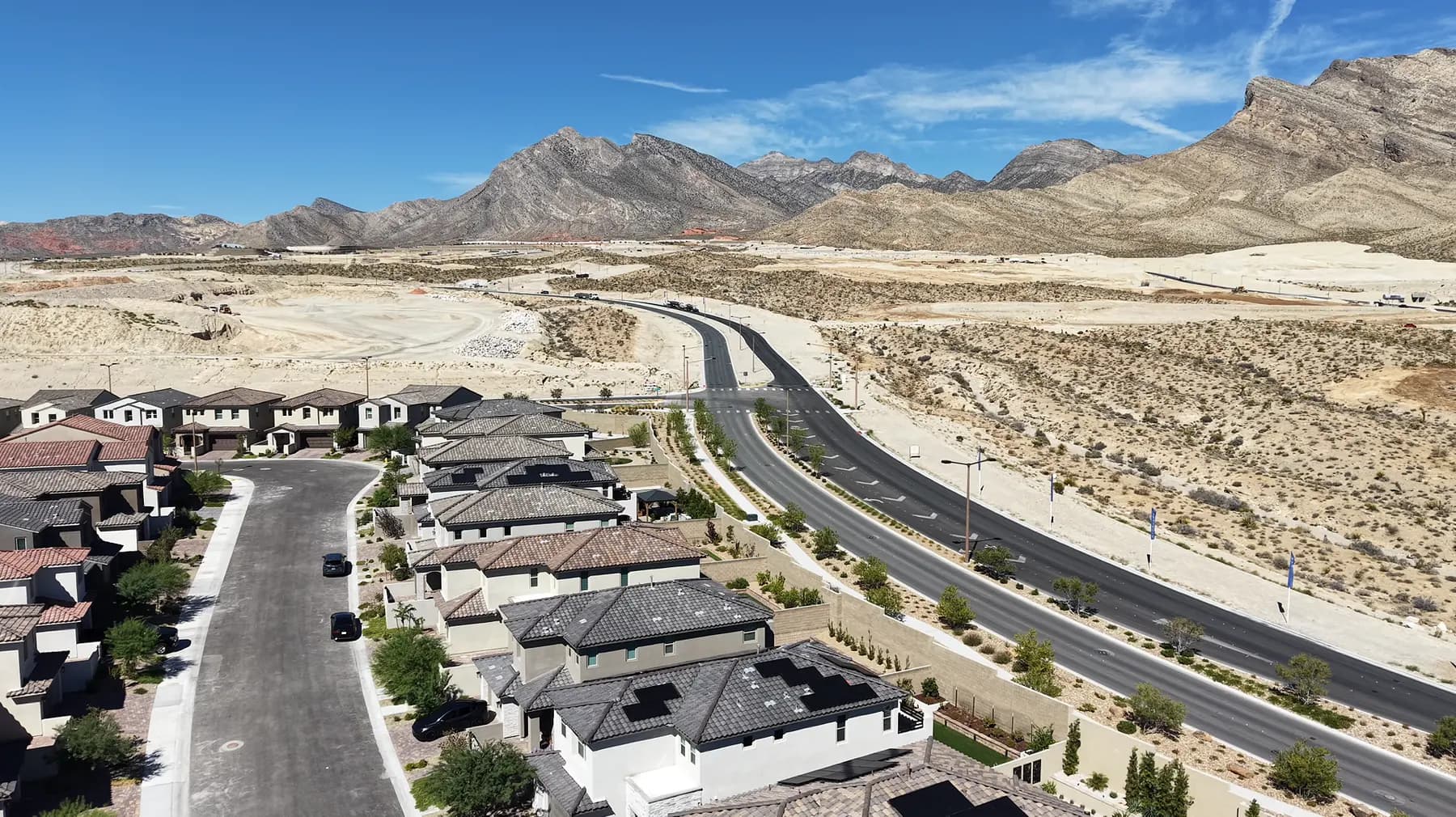 Aerial view of new homes at La Madre Peaks on the western edge of Summerlin, a wide arterial road running past graded desert toward the mountains.