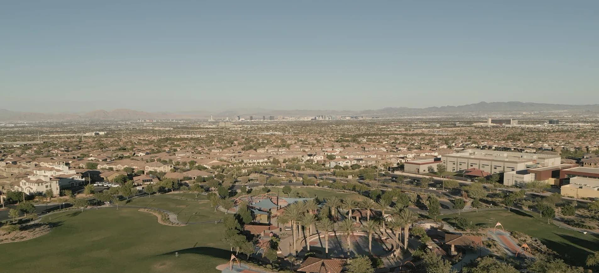 Aerial view looking east from above Mesa Ridge Park at the southern end of Summerlin, Las Vegas: a green park lawn with a playground, shaded ramadas and tall palms in the foreground, tile-roofed houses running to the horizon, the towers of the Las Vegas Strip small in the haze beyond, and desert mountain ranges on either side.
