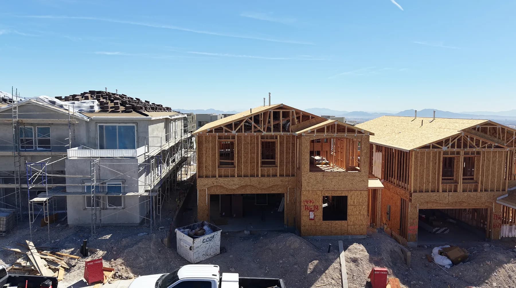New homes under construction in Summerlin West, open timber framing on two houses beside a finished stucco home still under scaffolding, with a dumpster and work truck at the curb.