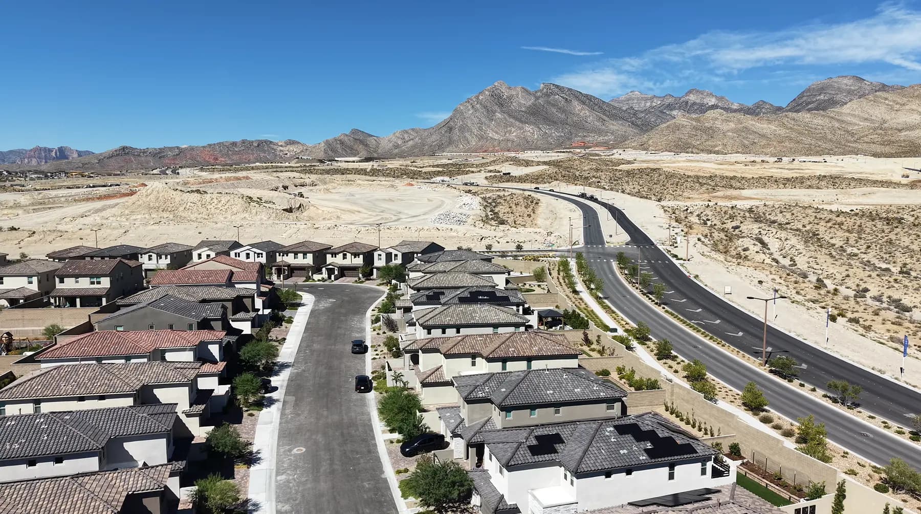 Aerial drone view of a new Summerlin West neighborhood: finished tile-roofed homes along a completed street on the left, a wide new arterial road on the right, and graded dirt pads and untouched desert running toward the mountains beyond.
