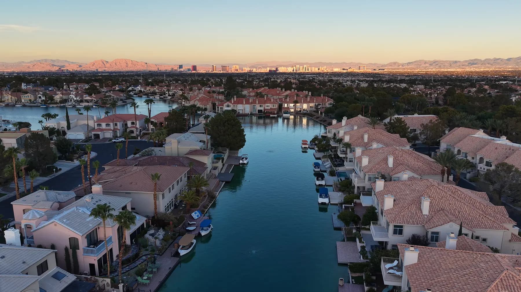 Aerial drone view at dusk over The Lakes in Las Vegas, an established waterfront neighborhood of tile-roofed homes with mature trees, private docks and small boats along the water, with the valley and the Strip skyline in the distance.