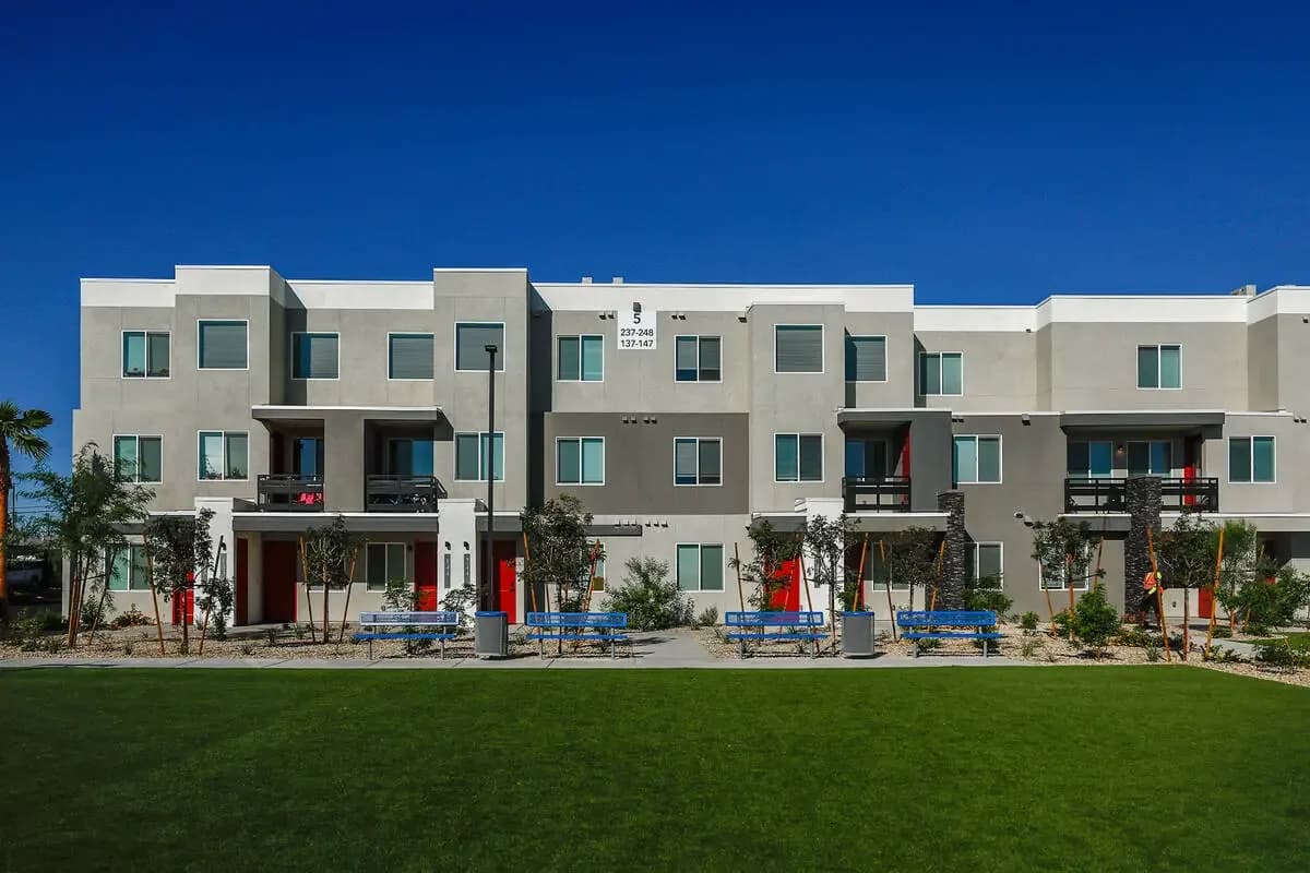 Straight-on view of a three-story stucco apartment building at Gholson Landing with red front doors, private balconies, new landscaping, blue benches, and a lawn in the foreground