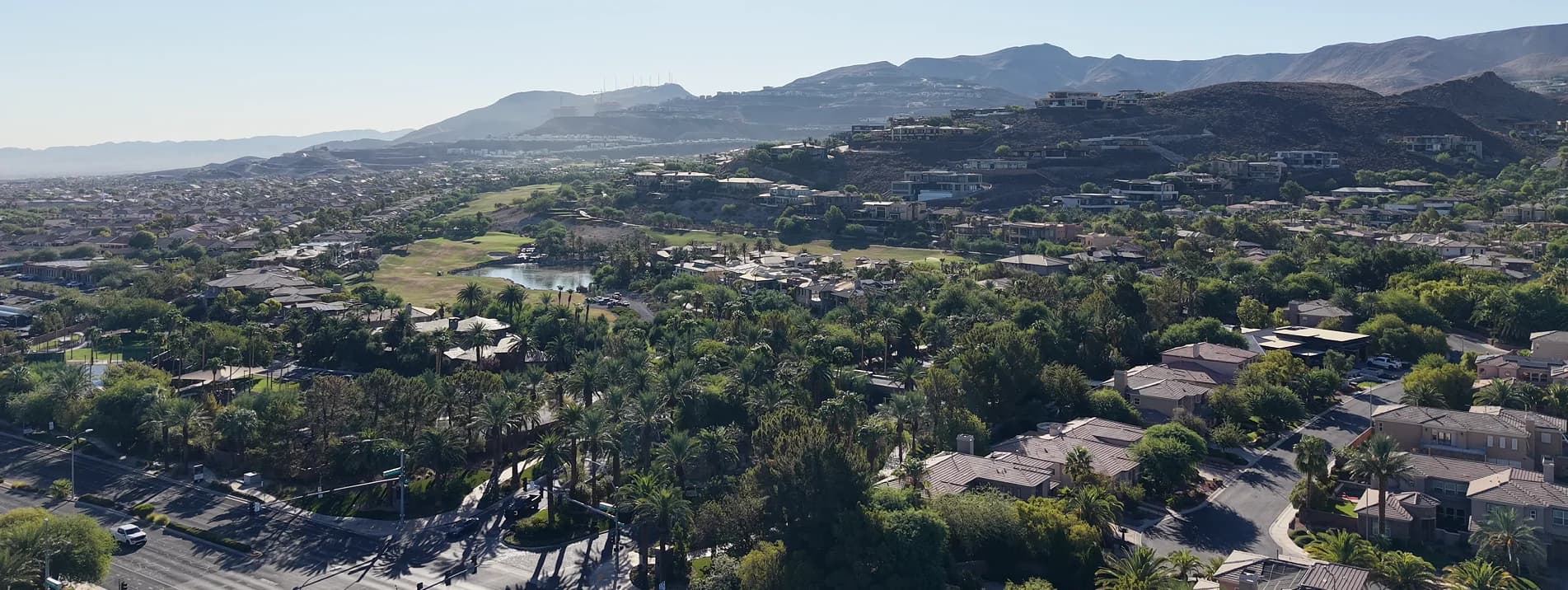 MacDonald Highlands, Henderson. Photo by Mikey Del Rosario Aerial view over Henderson, Nevada looking toward MacDonald Highlands: contemporary flat-roofed homes stepping up a dark rocky ridge in the middle distance, a golf course with a small lake below them, streets of tile-roofed houses under mature palms across the valley floor in the foreground, and the McCullough Range along the horizon.
