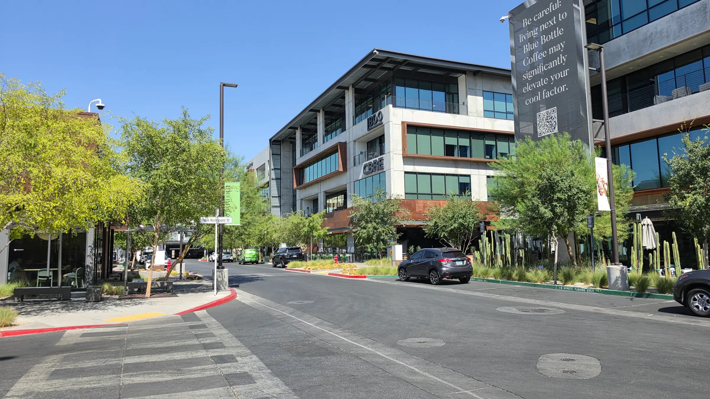 A quiet street through the UnCommons mixed-use campus in southwest Las Vegas at mid-morning, with three- and four-story offices housing BDO and CBRE, young shade trees, and desert landscaping along the curb.