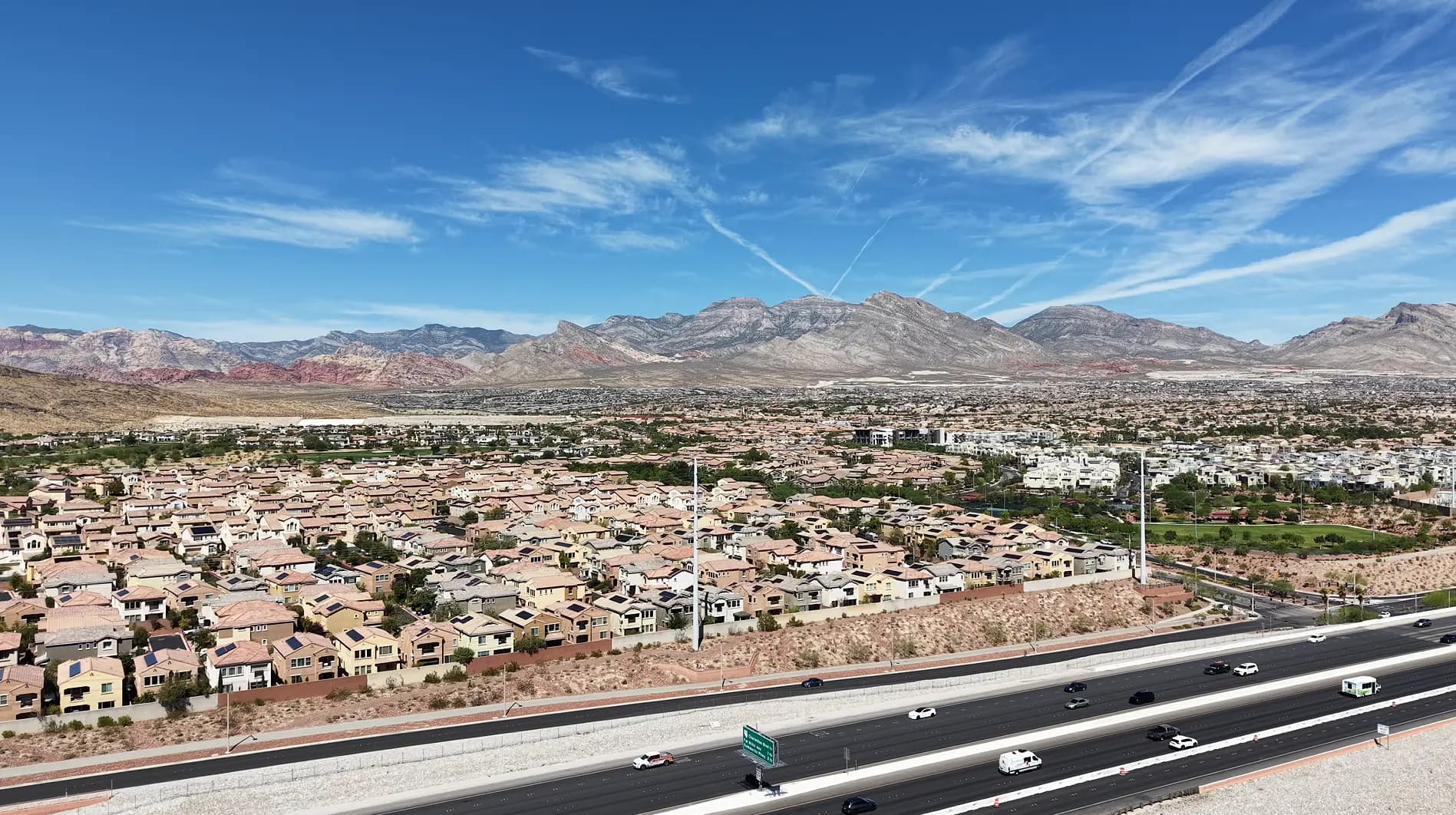 Aerial drone view over an established Las Vegas neighborhood of tile-roofed homes with grown-in trees and a green golf corridor, the 215 Beltway running across the foreground and the Red Rock escarpment and La Madre range on the horizon.