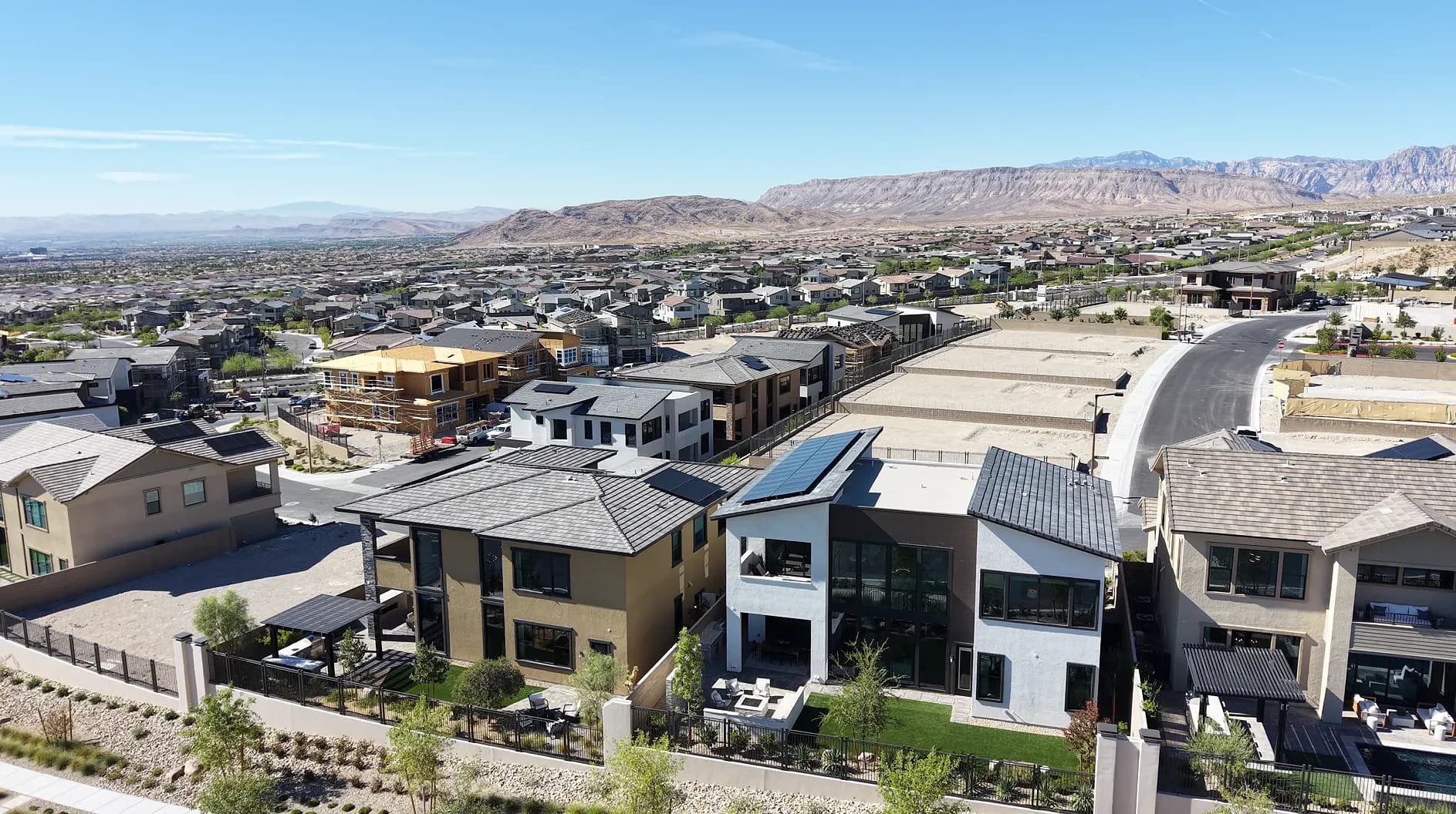 Aerial drone view over newer homes on the west side of Summerlin in Las Vegas: finished modern homes with rooftop solar in the foreground, homes still under construction behind them, and established neighborhoods running toward the Red Rock escarpment.