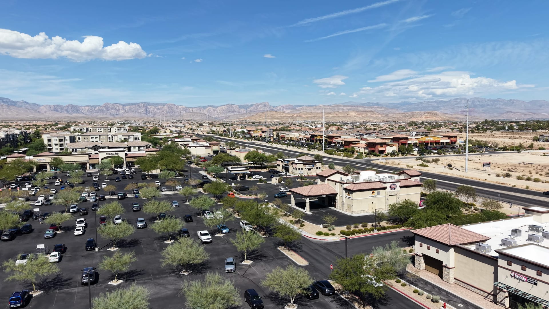 Aerial of a shopping center and wide road in southwest Las Vegas with neighborhoods and mountains in the distance.
