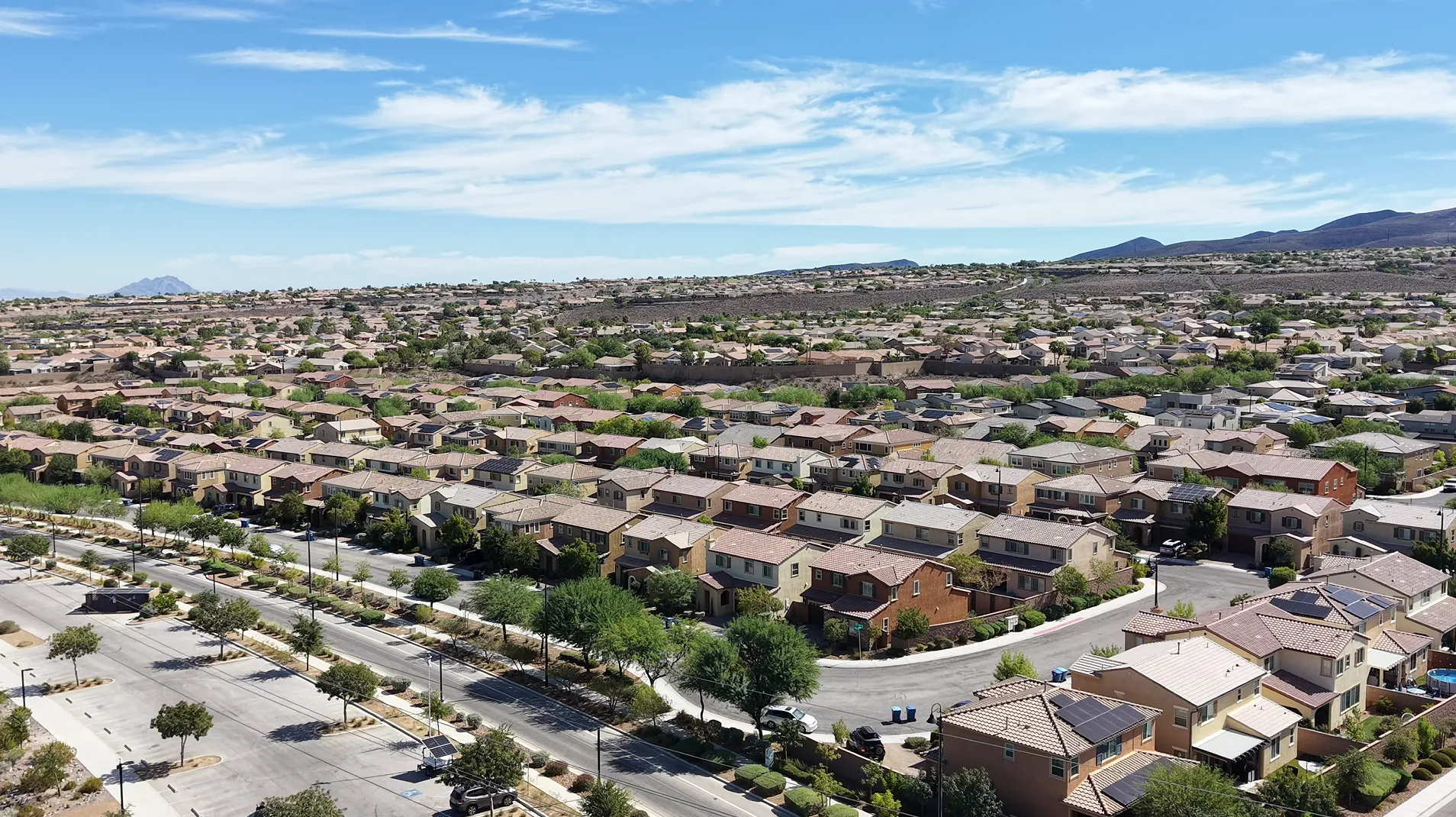 Aerial drone view of a Las Vegas residential neighborhood, with rows of tile-roofed tract homes, rooftop solar panels, and desert mountains under a blue sky in the background.