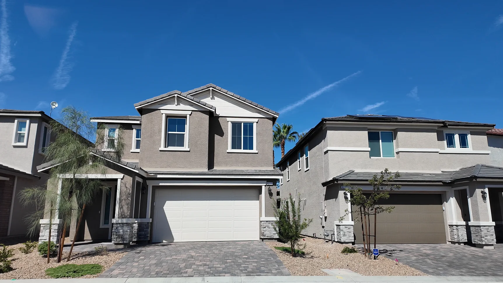 Two adjacent two-story stucco homes in a new Las Vegas subdivision, with tile roofs, rooftop solar on one home, desert rock landscaping, and small builder yard signs, under a clear blue sky.