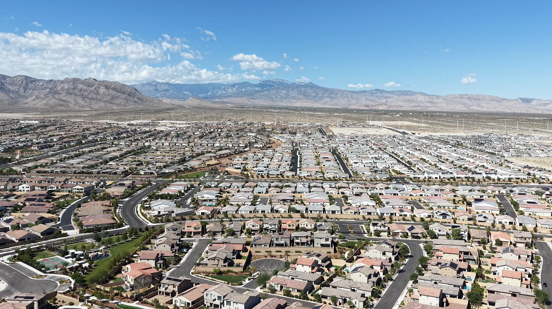 Aerial drone view of a Las Vegas suburban neighborhood: rows of tile-roofed homes and curving streets in the foreground, open desert beyond, and a mountain range under a blue sky with scattered clouds.