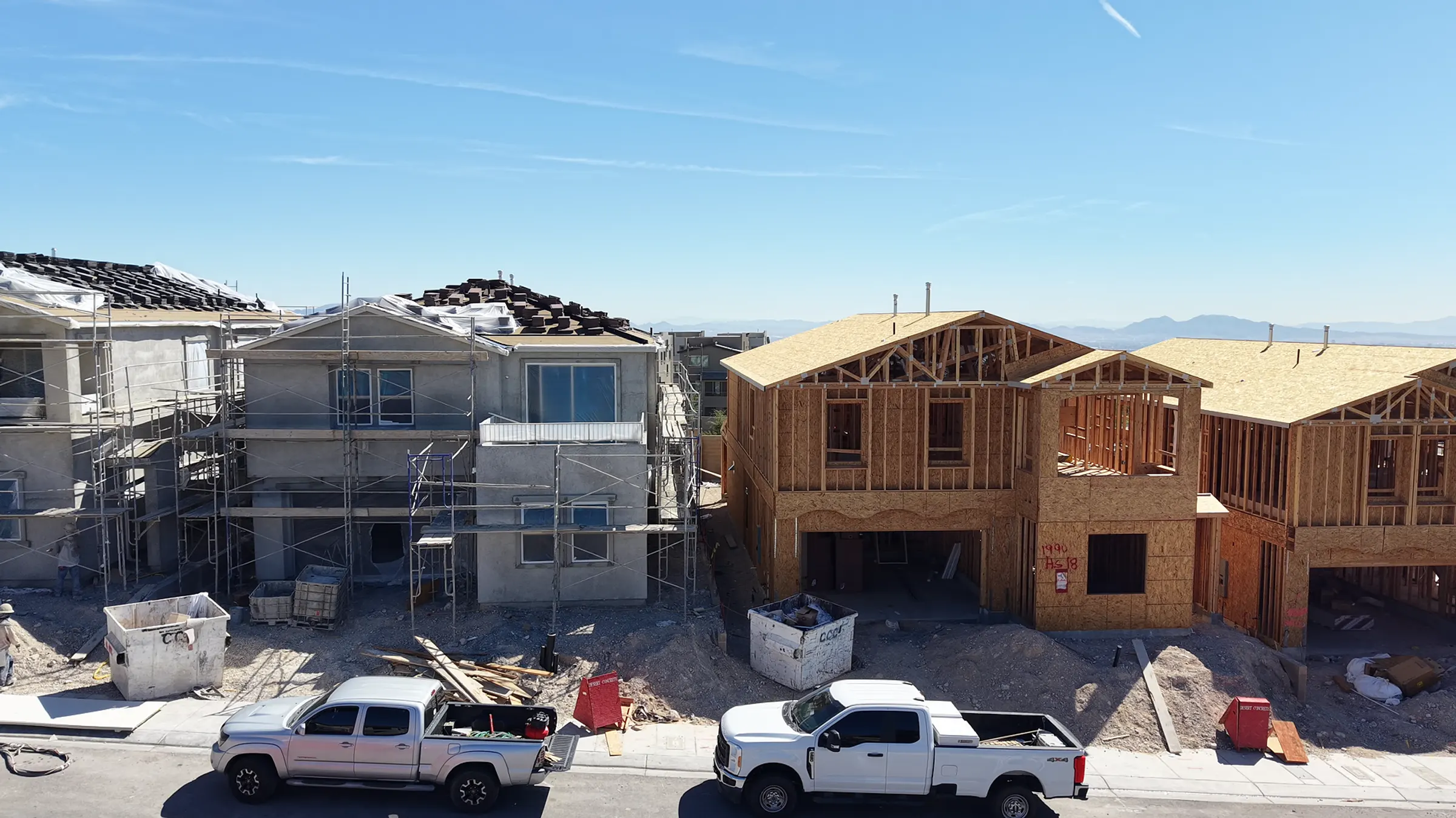 Elevated view of a Lennar new-construction street at Mockingbird in Summerlin, Las Vegas: finished stucco homes under scaffolding on the left, open timber framing mid-build on the right, work trucks and dumpsters along the curb, and the valley and mountains visible in the haze behind.