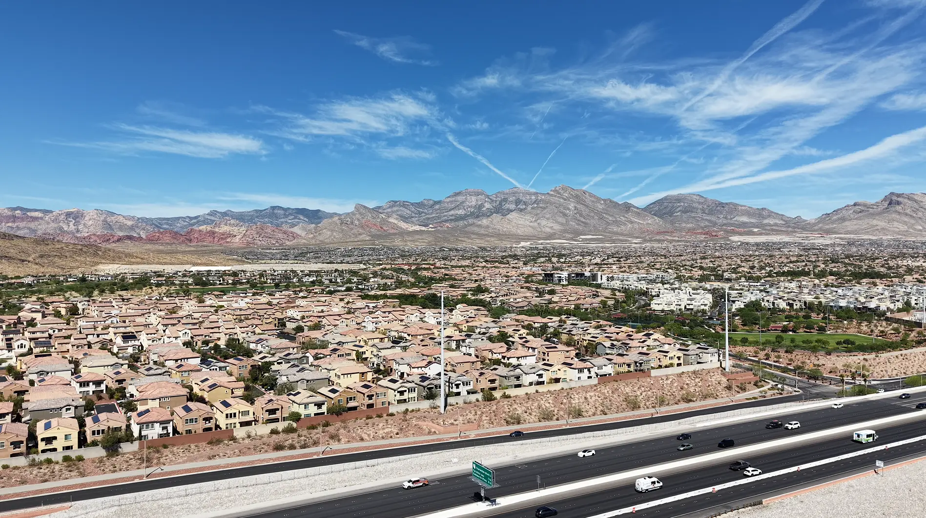 Aerial drone view over an established Las Vegas neighborhood of tile-roofed homes with grown-in trees and a green golf corridor, the 215 Beltway running across the foreground and the Red Rock escarpment and La Madre range on the horizon.