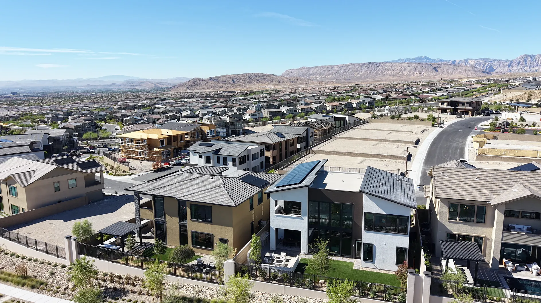 Aerial drone view over newer homes on the west side of Summerlin in Las Vegas: finished modern homes with rooftop solar in the foreground, homes still under construction behind them, and established neighborhoods running toward the Red Rock escarpment.